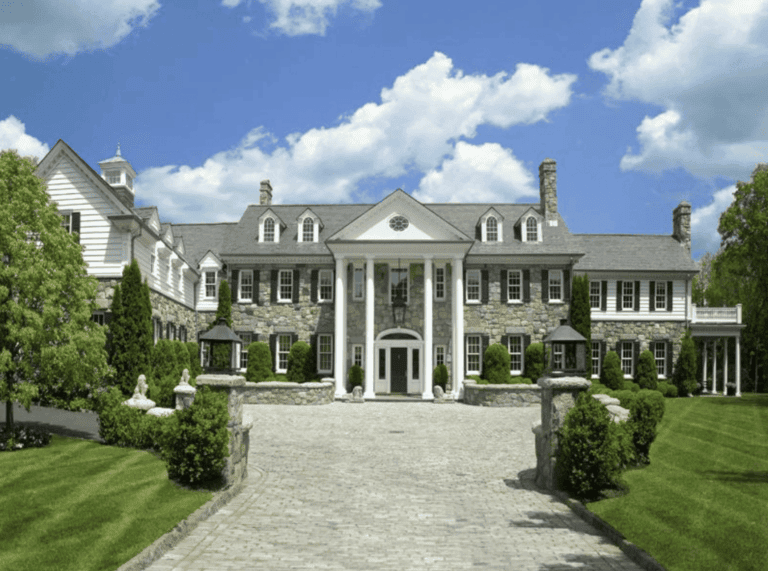 Luxury colonial mansion with stone and white siding, grand front entrance with tall columns, manicured lawn, lush greenery, blue sky with clouds, elegant residential architecture for estate living, Iridium Development Inc.