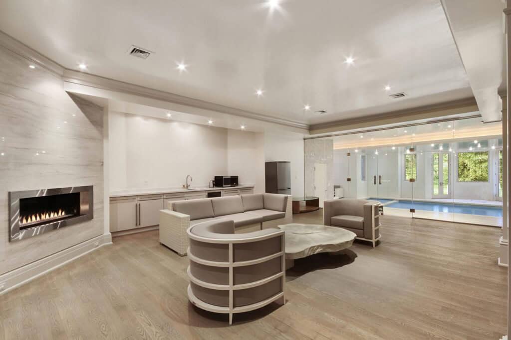 Modern luxurious indoor lounge area with fireplace, neutral-toned furniture, and large glass pool door, showcasing elegant interior design and advanced home automation features.