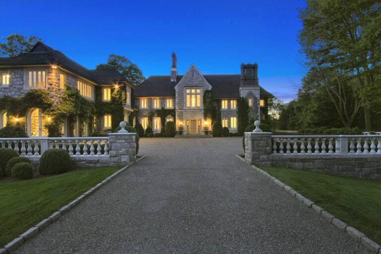 Luxurious stone mansion with illuminated windows, elegant driveway, lush garden, and timeless architecture, showcasing upscale residential design and landscaping.