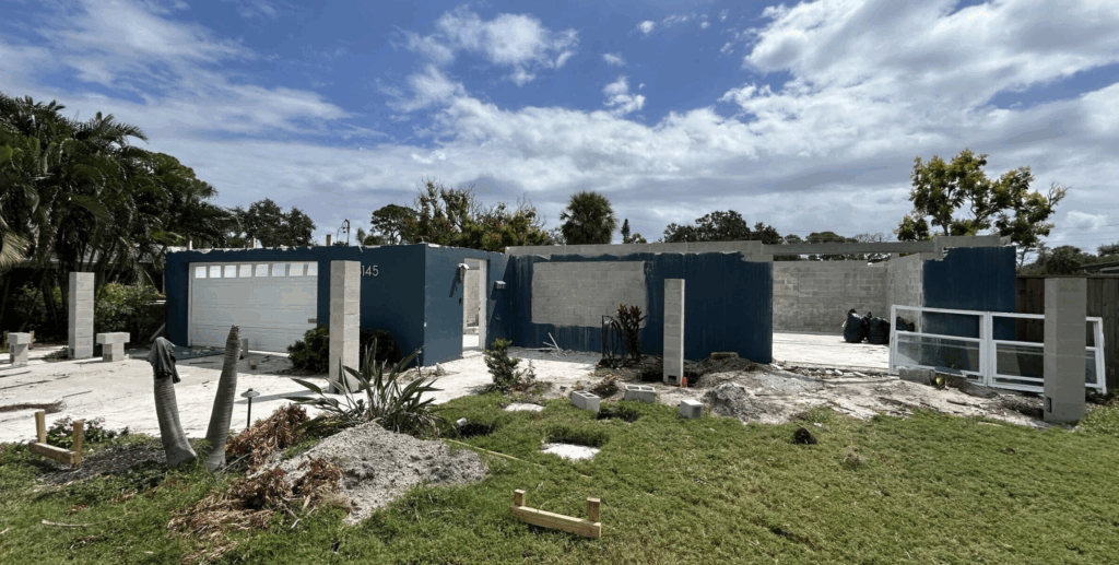 Modern house under construction with blue exterior walls, concrete blocks, and ongoing work, surrounded by greenery and trees, in a sunny setting with a partly cloudy sky.